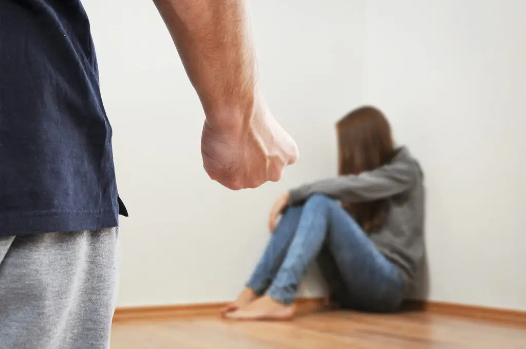 Man's clenched fist threateningly raised towards a woman sitting in a corner, illustrating the concept of domestic violence and its emotional impact.