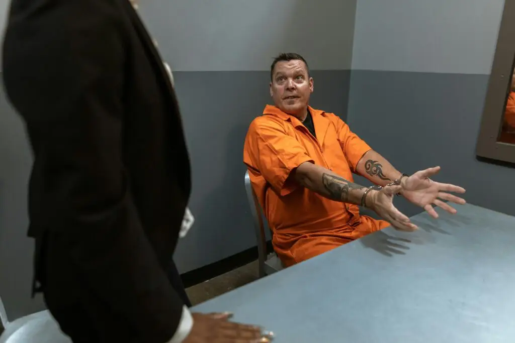 Man in orange prison jumpsuit with handcuffs gesturing during a legal consultation with an attorney in a formal suit, emphasizing the importance of legal representation in criminal defense.