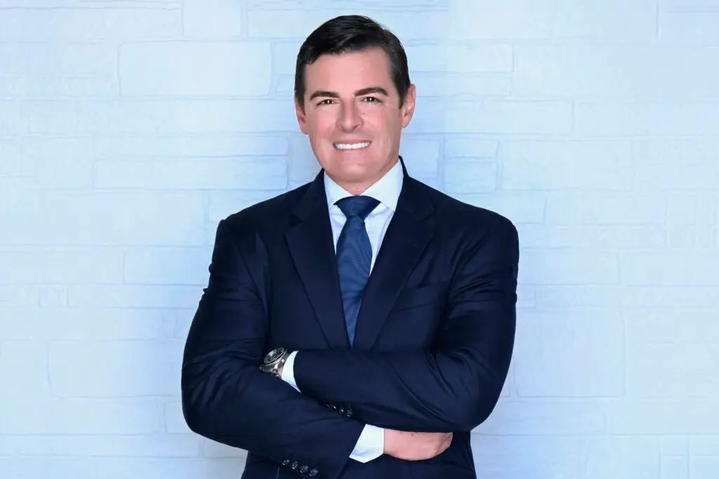 Broward County Criminal Defense Attorney Michael White in a navy suit standing against a light blue brick wall with arms crossed, dressed professionally in a white shirt and dark blue tie